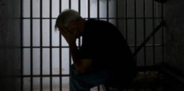 Silhouettee of a man sitting in a jail cell, holding his head in his hands, with cell bars behind him