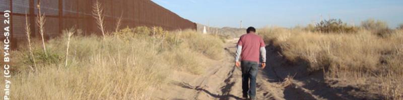 Photo of a man walking down a dirt road, away from the camera. He is surrounded by desert scrub, and on his left is the U.S.-Mexico border fence stretching to the horizon.