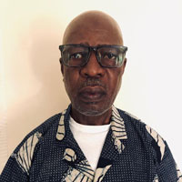 Photo of a man named Goura. He is looking at the camera with a somber expression. He wears glasses with thick dark rims and you can just see the blue collar of his shirt. The background is plain white.