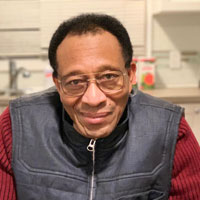 Photo of a man named Ibrahima. He is looking at the camera with a slight smile on his face. He wears glasses, has short dark hair, and is wearing a black leather zip-up vest over a red sweater. The background of the photo is his kitchen.
