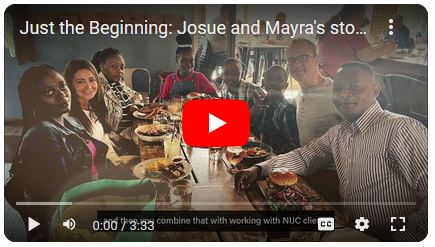 Still image of YouTube video with play icon. In video is a Haitian family sitting around a table together in a restaurant smiling and posing for a photo.