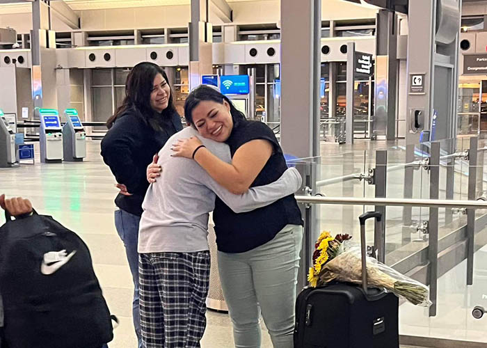 A Latina mother hugs her teenage son with a big smile on her face in an airport terminal after they have been separated for three years. Her daughter looks on in the background smiling widely.