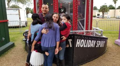 A mother and father hug their two teenage daughters. The mother holds her toddler son. They're standing outside in front of a holiday display.