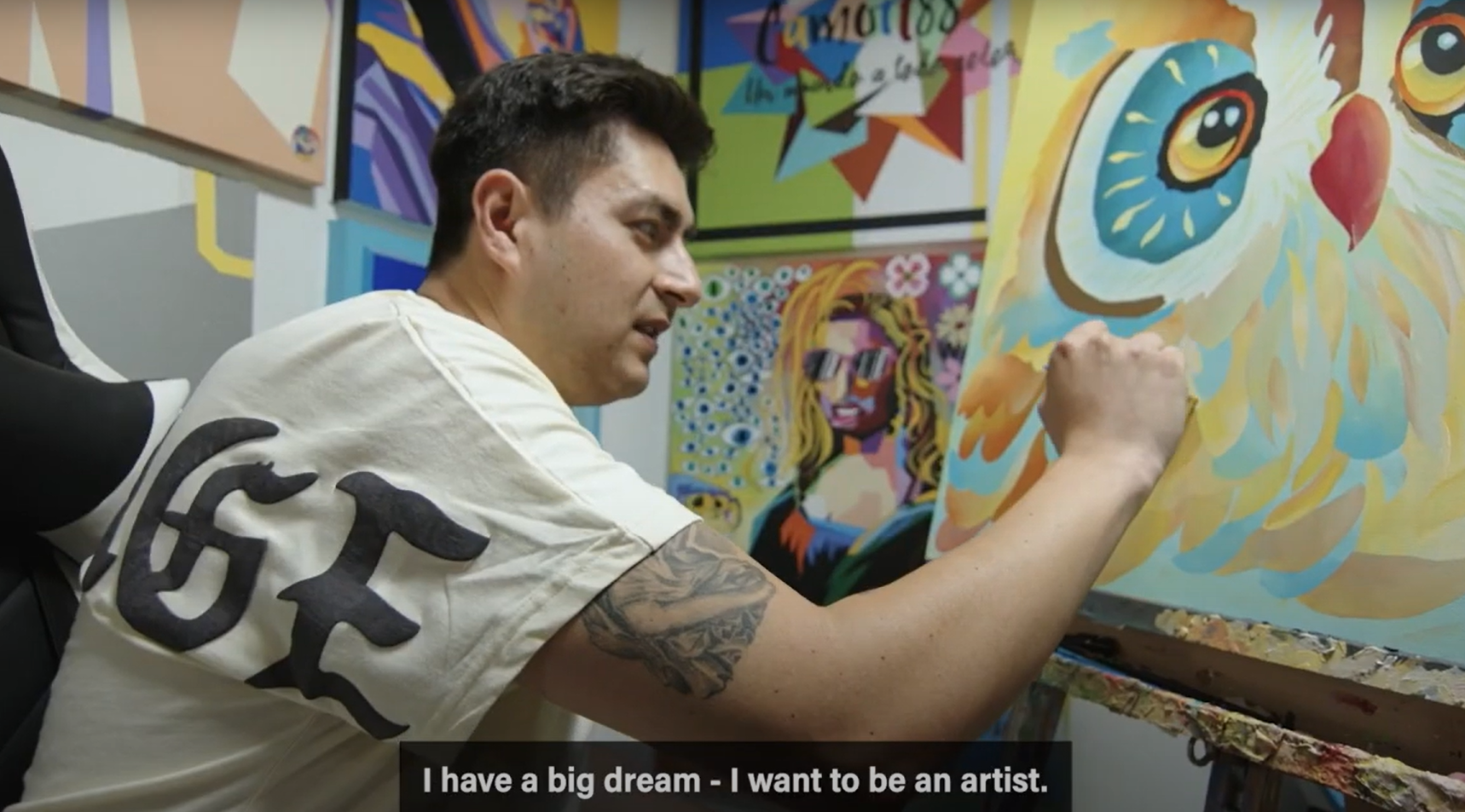 Still image of YouTube video. In video is NIJC client Camilo sitting at an artist's easel, paintbrush in hand, adding detail to a painting of an owl.