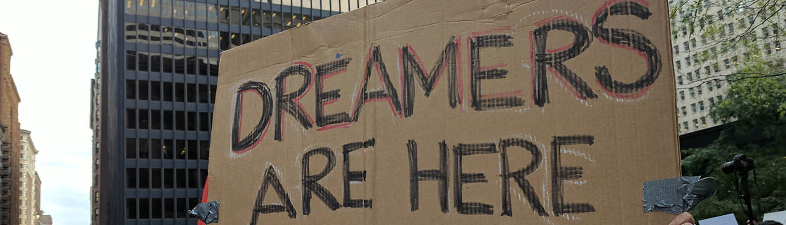 Mano con un cartel de protesta que dice "Los soñadores están aquí para quedarse" con los edificios de la ciudad al fondo