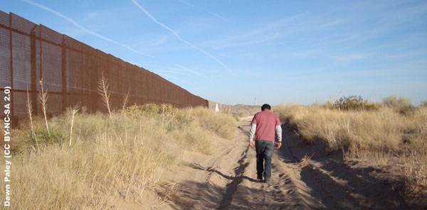 Photo of a man walking away from the camera along a dirt road, with desert scrub on either side. On his left is the US-Mexico border fence