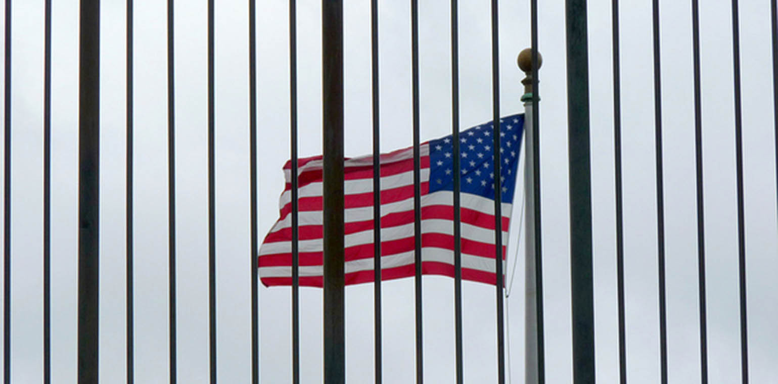 Photo of a U.S. flag flying toward the left, with a blue sky in the background and iron bars in the foreground.