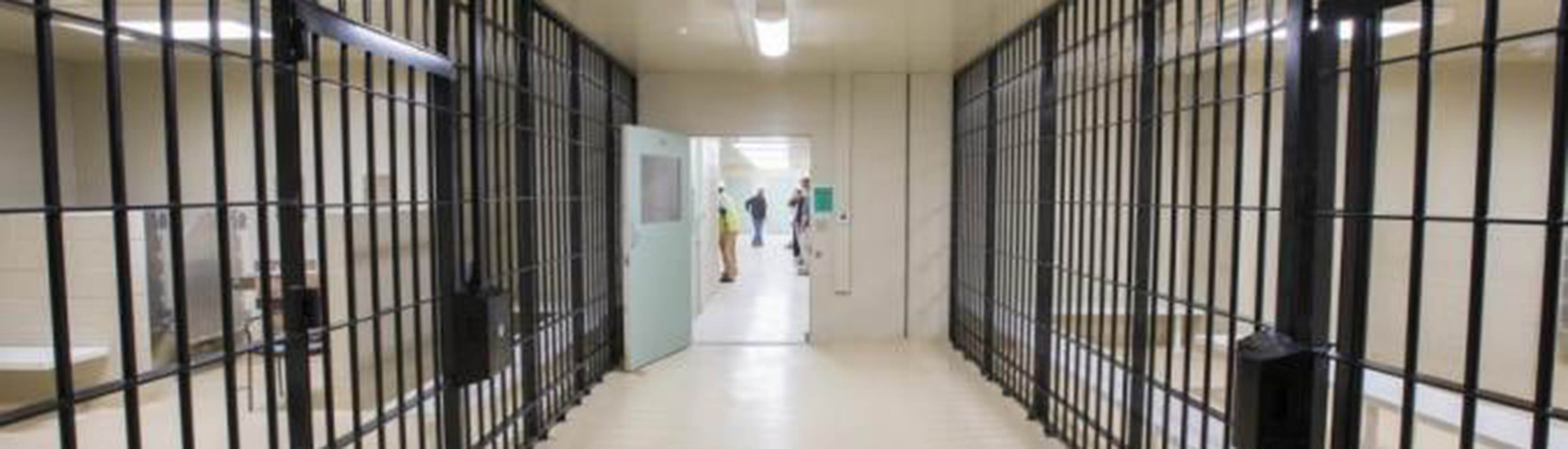Looking down a hallway in a jail. Cell bars line either side of the hallway. at the end of the hallway there is an open door where you can see people walking and standing.