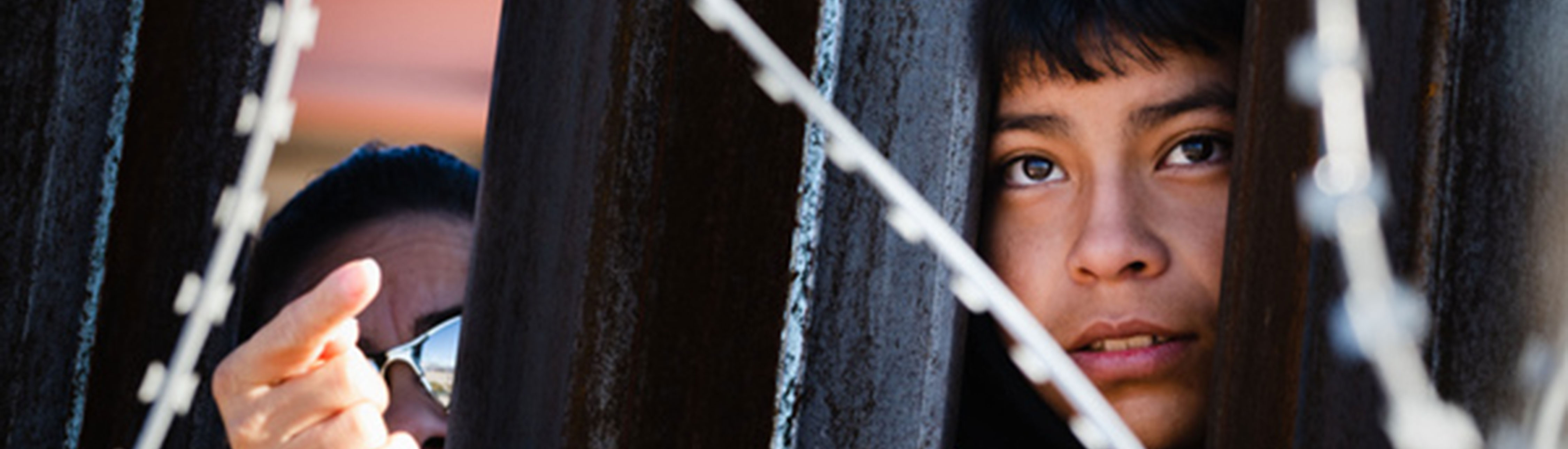 Photo of a boy's face looking between two iron bars of the southern border fence, with barbed wire in the foreground and another person's hand to his right pointing through the fence.