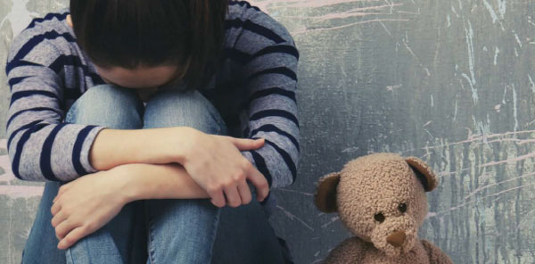 Image from the cover of the new report Punishing Trauma - a young child holds her head in her curled arms and knees, with a teddy bear by her side.