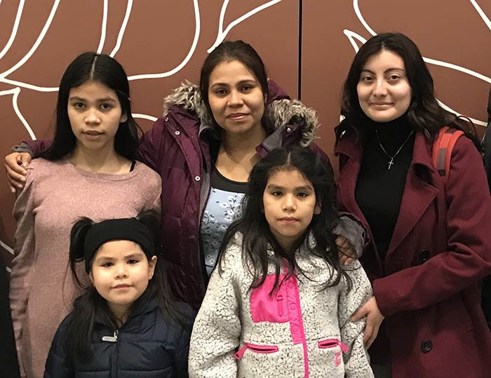 A family with two adults and three young girls stand in a law firm lobby and smile after winning asylum