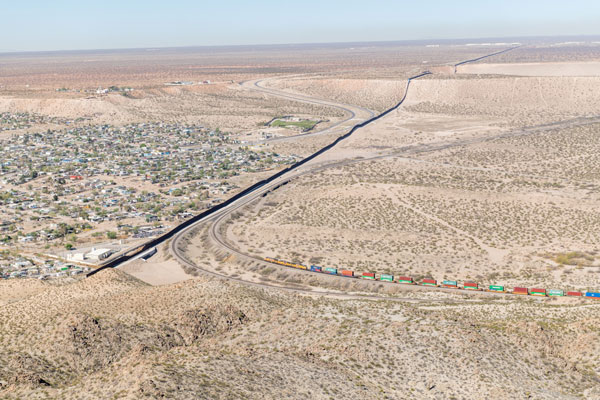 Photo of the border between Juarez, Mexico, and El Paso, Texas, from a hill overlooking El Paso. An iron fence cuts between the two, with the city of Juarez on one side and a desert with a train track on the other.