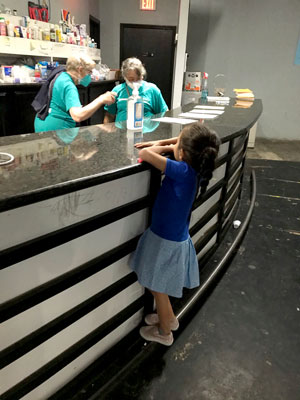 Photo of a pharmacy counter staffed by two pharmacists working. A young girl in blue, with her back to the camera, stands on her tiptoes looking over the counter.