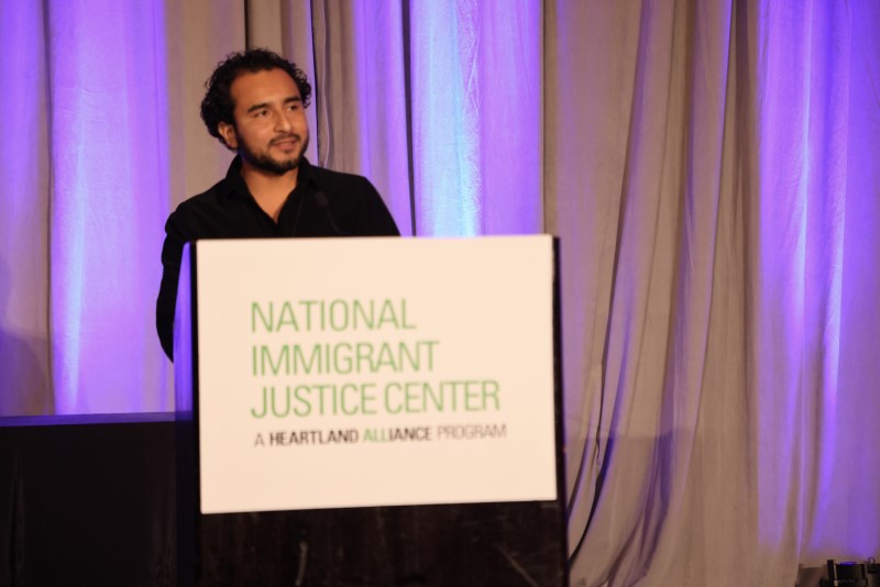 Javier Zamora, a poet and author, looks pensive as he delivers the keynote address from a podium with a sign on it with the "National Immigrant Justice Center" logo