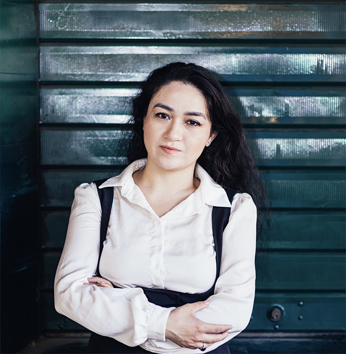 headshot of woman in a white button down shirt posing in front of a stark black background. She has long curly dark hair and looks confidently and deeply into the camera.  