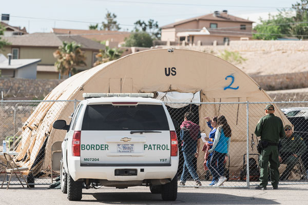 A photo of a Border Patrol truck parked next to a chainlink fence. On the other side of the fence is a large arched tent with border agents and three people standing near the entrance.