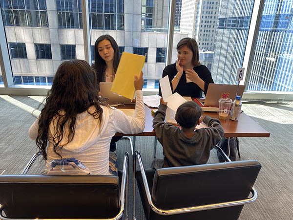 Two women volunteer lawyers sit at a table meeting with a mother and her young son in an office with huge windows and a skyscraper behind them. They look a paperwork together.