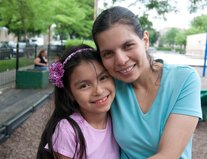 A Latina mother hugs her young daughter tightly, both are smiling widely