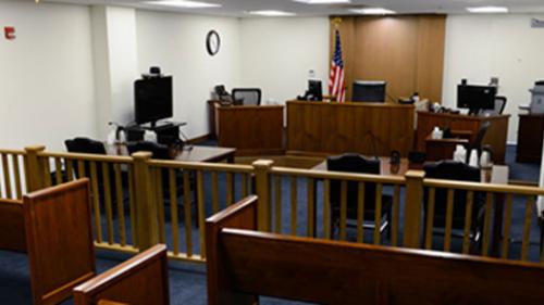 Photo of inside an immigration court room. A room with white walls and a blue carpet, a wooden fence down the middle with wooden benches on one side and two tables and a judge's bench on the other.