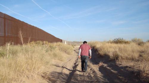 Photo of a man walking down a dirt road, away from the camera. He is surrounded by desert scrub, and on his left is the U.S.-Mexico border fence stretching to the horizon.