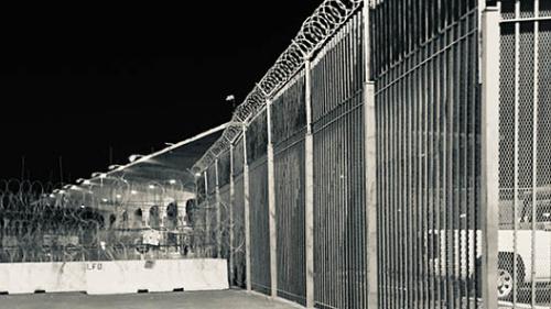 A nighttime photo of a high fence topped with barbed wire, at a militarized checkpoint at the U.S.-Mexico border.
