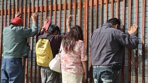 People face the border wall at the U.S.-Mexico border, with their hands up against the wall. Border agents with guns stand by.