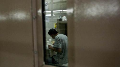 A photo taken through the narrow window of a heavy prison door. Through the window you see the profile of a man, angled away from the camera so you cannot see his face, sitting bent over a table, wearing an jail-issued uniform.