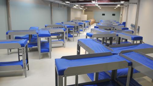 Photo of a large dorm room inside Farmville ICA detention center. Bunk beds with blue mattresses are arranged in groups of four inside a large room with bright fluorescent lighting and concrete walls  