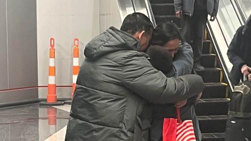 Photo of a mother and father embracing their son, all wearing winter coats, standing next to an airport escalator
