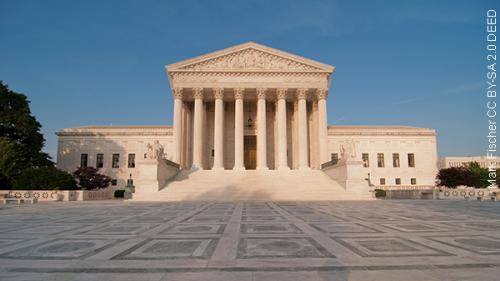 A photo with the U.S. Supreme Court building in the center of the image, the concrete plaza stretching in front of the building toward the foreground and a clear blue sky in the background.