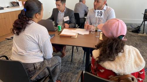 Two attorneys sit across a small table from two women. One attorney is typing on the computer and the other is talking to the women about their legal case.