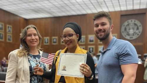 Photo of NIJC client Karima at her U.S. citizenship oath ceremony, holding a small U.S. flag in her right hand and her naturalization certificate in her left hand. To her right and left are pro bono attorneys who assisted in her case.