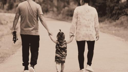 A toddler walks in between their mother and father, reaching up to grab their hands as they walk along a tree-lined path.