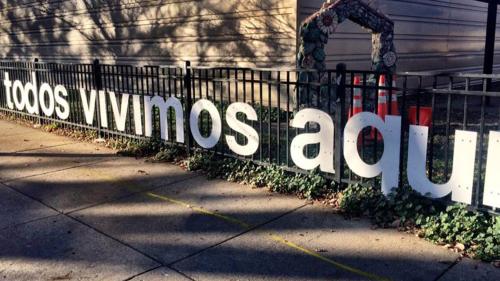 Large white letters line a black fence outside of a school reading, "todos vivimos aqui."