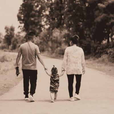 A toddler walks in between their parents, reaching up to grab their hands as they walk along a tree lined path