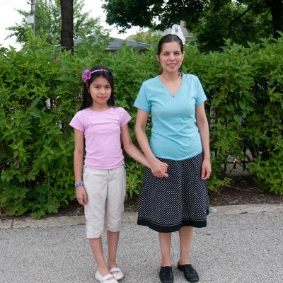 A mother and young daughter stand together holding hands. They are outside on a warm summer day and are standing in front of green leafy bushes. 