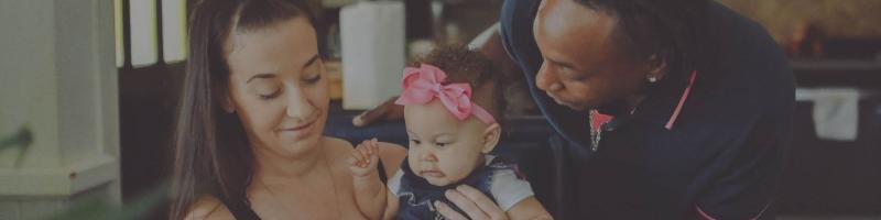 A mother holds a baby on her lap while the father looks down adoringly.