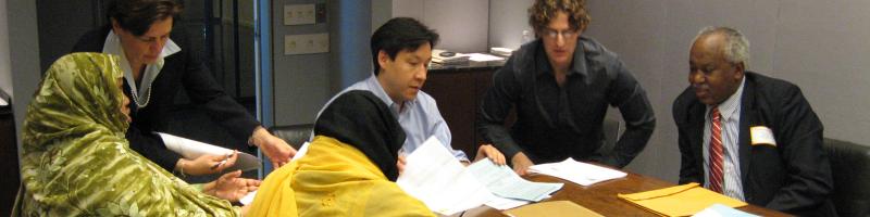Several men and women lawyers sit at a table in an office and sort through a pile of papers. Also at the table are two women in headscarves who have documents and are filling out forms.