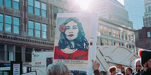Photo of a group of people at a rally in a city downtown area. A person with their back to the camera is holding up a poster with an illustration of a woman with long dark hair, red dress, and flower in her hair above the phrase "We The People."