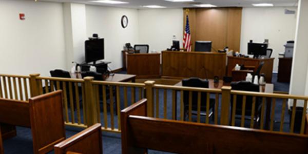 Photo of inside an immigration court room. A room with white walls and a blue carpet, a wooden fence down the middle with wooden benches on one side and two tables and a judge's bench on the other.