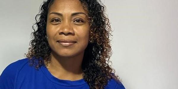 Close-up photo of a woman named Assia, who is smiling at the camera. Her face is framed by curly brown hair, and she is wearing a bright blue shirt. The background is plain white.