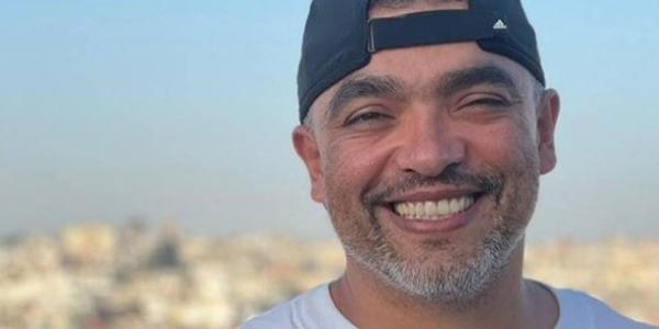 Close-up photo of Carlos smiling at the camera. He is wearing a baseball cap backwards and white t-shirt. In the background is a blue sky and blurred overhead view of buildings and trees.