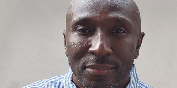 Close-up photo of the face of Samuel, who is looking calmly at the camera. He is wearing a collared shirt that is just visible, and the background is plain white.