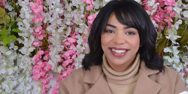 Photo of a woman named Tina, who is smiling at the camera. She is wearing a tan blazer and turtle neck and has a sleek short bob haircut with bangs. Pink, white, and green flower garlands make up the background.