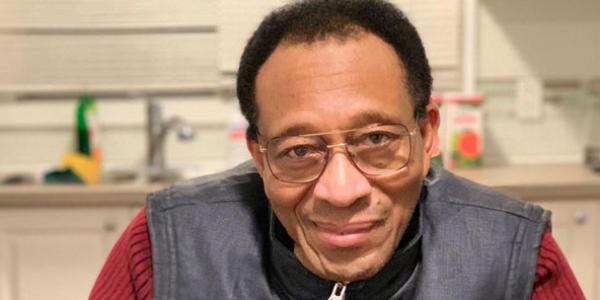 Photo of a man named Ibrahima. He is looking at the camera with a slight smile on his face. He wears glasses, has short dark hair, and is wearing a black leather zip-up vest over a red sweater. The background of the photo is his kitchen.