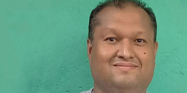 Photo of Juan Carlos, looking at the camera with a calm smile. He has close-cropped dark hair and is wearing a gray shirt and a cross on a cord around his neck. The background is bright teal green.