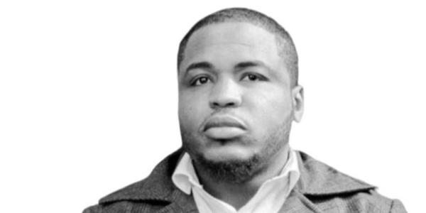 Close-up black and white headshot of Paul, who is looking at the camera with a neutral expression. He is wearing a wide-collared jacket over a light-colored collared shirt. The background is white.