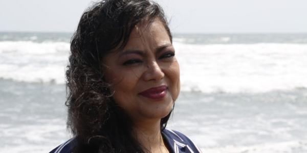 Close-up headshot photo of a woman named Vanessa. She is standing on the beach and looking at the camera with a slight smile. She has long dark hair and a black and white collared shirt on. In the background you can see the ocean waves.