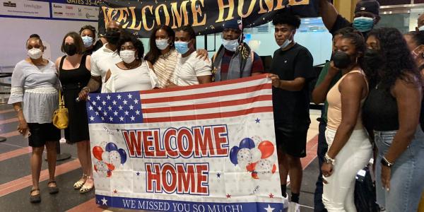 A group of men 14 men and women gather together holding two "Welcome Home" banners. One of the flags is decorated with the U.S. flag and also states "We Missed You So Much"