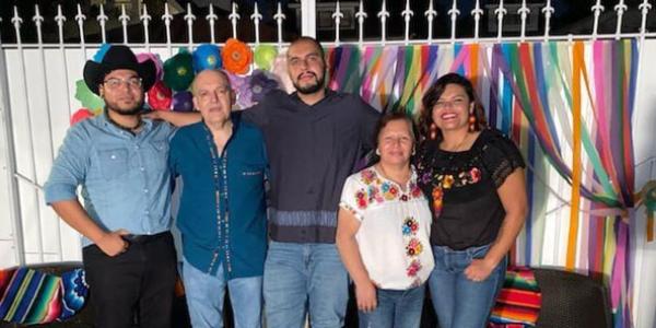 Beto stands in the center of a group of five smiling family members, in front of a white fence decorated with multi-colored streamers.